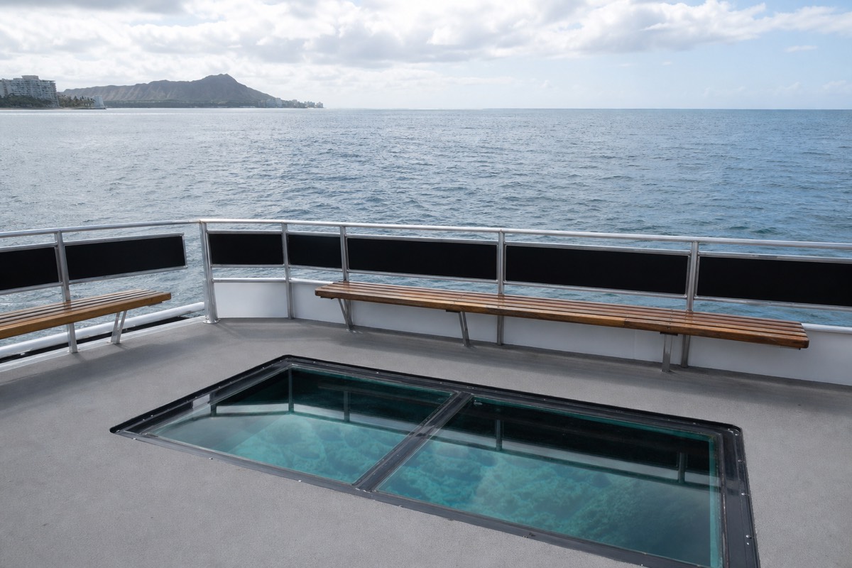 Passengers looking down through a glass viewport at a coral reef from the deck of a glass-bottom boat off Oahu